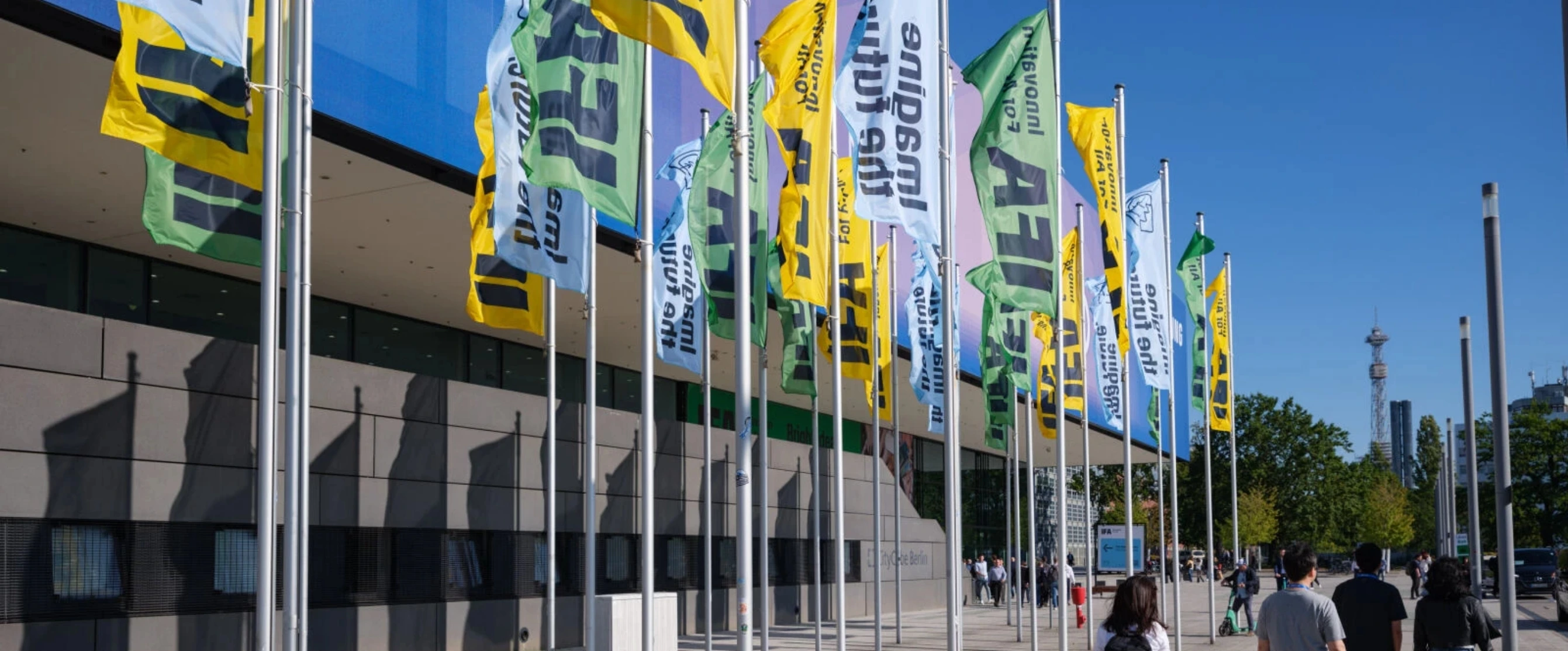 Reihe bunter IFA-Fahnen (gelb, grün, blau) vor Messegebäude bei klarem Himmel, Besucher im Vordergrund