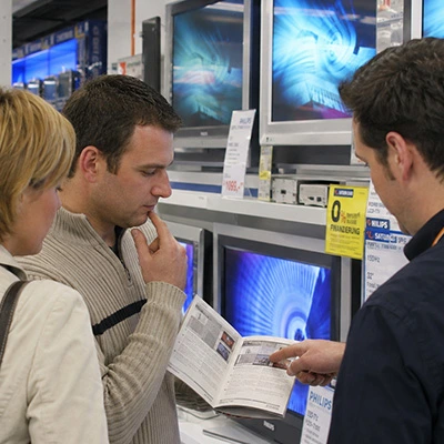Kundengespräch 2006: Zwei Kunden vergleichen Fernseher im Elektronikmarkt, Verkäufer zeigt Prospekt und Modelle