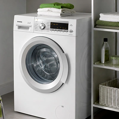 Weiße Frontlader-Waschmaschine (Siemens) in Waschküche mit gefalteten Handtüchern und Regal mit Waschmittel