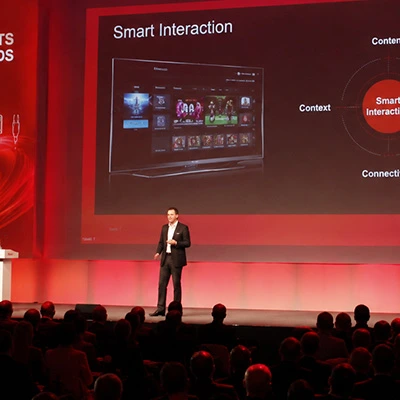 Präsentation 2012: Redner auf roter Bühne vor Publikum zeigt Smart-Interaction-Bildschirm mit TV-Oberfläche und Grafiken