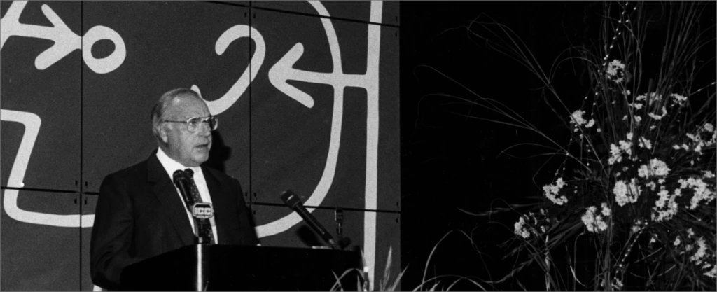 Schwarzweiß-Foto: Redner am Mikrofon auf IFA-Bühne neben Logo und Blumenarrangement, Historie 1980er