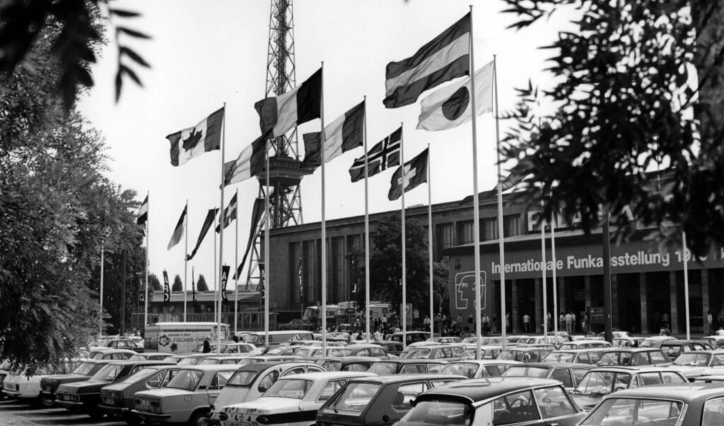 Schwarzweiß-Foto: voller Parkplatz vor Messegebäude mit Flaggen verschiedener Länder bei historischer IFA-Funkausstellung