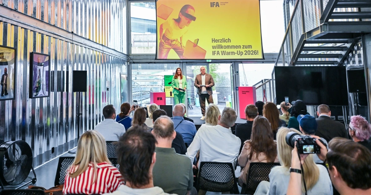 Publikum schaut auf Bühne beim IFA Warm‑Up 2026: Moderatoren präsentieren vor großem Bildschirm und Messestand in Halle
